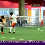 Dois jogadores disputam uma partida de futebol de cegos em quadra coberta. Um deles, com uniforme preto do Corinthians, domina a bola enquanto o adversário, de uniforme branco e shorts laranja, tenta a marcação. Ao fundo, pessoas assistem atrás da grade de proteção. Na parte inferior da imagem, legenda do programa Globo Esporte: ‘Corinthians tem agora futebol de cegos e disputa a Liga Nacional’.