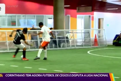Dois jogadores disputam uma partida de futebol de cegos em quadra coberta. Um deles, com uniforme preto do Corinthians, domina a bola enquanto o adversário, de uniforme branco e shorts laranja, tenta a marcação. Ao fundo, pessoas assistem atrás da grade de proteção. Na parte inferior da imagem, legenda do programa Globo Esporte: ‘Corinthians tem agora futebol de cegos e disputa a Liga Nacional’.