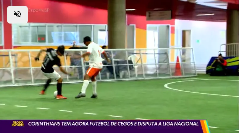 Dois jogadores disputam uma partida de futebol de cegos em quadra coberta. Um deles, com uniforme preto do Corinthians, domina a bola enquanto o adversário, de uniforme branco e shorts laranja, tenta a marcação. Ao fundo, pessoas assistem atrás da grade de proteção. Na parte inferior da imagem, legenda do programa Globo Esporte: ‘Corinthians tem agora futebol de cegos e disputa a Liga Nacional’.