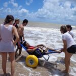 Jovem com deficiência é levado ao mar em uma cadeira anfíbia com rodas grandes e amarelas, acompanhado por um grupo de pessoas que o auxiliam. Eles estão na beira da praia, com ondas suaves e céu azul ao fundo.