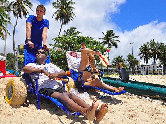 Homem com deficiência sentado em cadeira de rodas adaptada para areia, sendo empurrado por outro homem em uma praia ensolarada. Ao lado, uma mulher também relaxa em cadeira adaptada, enquanto pessoas aproveitam o dia próximo ao mar. Ao fundo, coqueiros e prédios completam a paisagem.