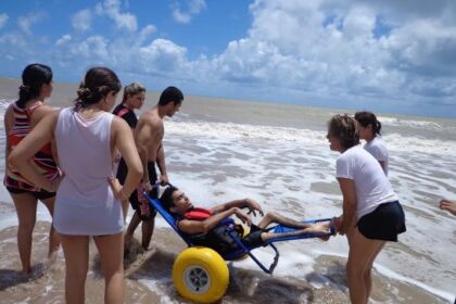 Jovem com deficiência é levado ao mar em uma cadeira anfíbia com rodas grandes e amarelas, acompanhado por um grupo de pessoas que o auxiliam. Eles estão na beira da praia, com ondas suaves e céu azul ao fundo.