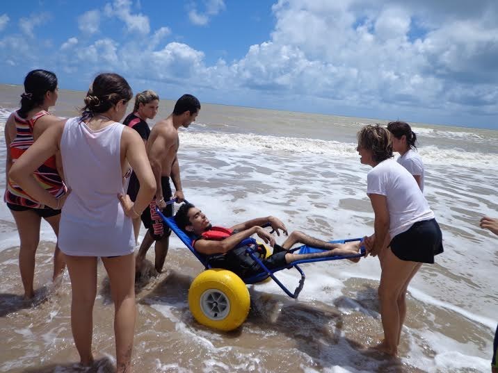 Jovem com deficiência é levado ao mar em uma cadeira anfíbia com rodas grandes e amarelas, acompanhado por um grupo de pessoas que o auxiliam. Eles estão na beira da praia, com ondas suaves e céu azul ao fundo.