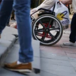 Pessoa em cadeira de rodas tenta transitar por calçada com desnível e degraus, enquanto outra pessoa empurra a cadeira. Um pedestre sobe a escada ao lado, mostrando a dificuldade de acessibilidade urbana.