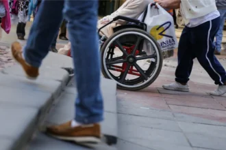 Pessoa em cadeira de rodas tenta transitar por calçada com desnível e degraus, enquanto outra pessoa empurra a cadeira. Um pedestre sobe a escada ao lado, mostrando a dificuldade de acessibilidade urbana.