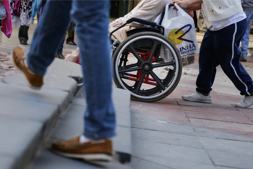 Pessoa em cadeira de rodas tenta transitar por calçada com desnível e degraus, enquanto outra pessoa empurra a cadeira. Um pedestre sobe a escada ao lado, mostrando a dificuldade de acessibilidade urbana.
