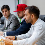 Deputados distritais em uma mesa de conferência, com a atenção voltada para um homem barbudo, de camisa branca e brinco na orelha, no primeiro plano. Ele está segurando alguns papéis e falando em um microfone que está à sua frente. Ao lado dele, há um homem usando um boné vermelho, e mais ao fundo, outras pessoas desfocadas