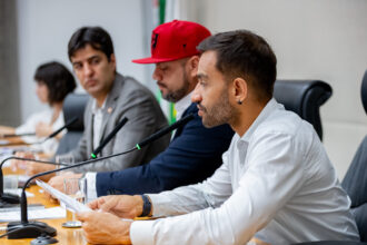 Deputados distritais em uma mesa de conferência, com a atenção voltada para um homem barbudo, de camisa branca e brinco na orelha, no primeiro plano. Ele está segurando alguns papéis e falando em um microfone que está à sua frente. Ao lado dele, há um homem usando um boné vermelho, e mais ao fundo, outras pessoas desfocadas