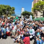 Uma grande foto de grupo tirada ao ar livre, em um dia ensolarado, mostrando dezenas de pessoas participando da 2ª Caminhada da Pessoa com Deficiência. O grupo é diverso e inclui pessoas com e sem deficiência, adultos e crianças, alguns em cadeiras de rodas e alguns usando camisetas brancas com um logo estampado.