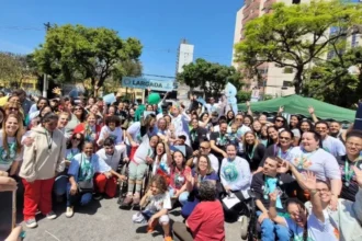 Uma grande foto de grupo tirada ao ar livre, em um dia ensolarado, mostrando dezenas de pessoas participando da 2ª Caminhada da Pessoa com Deficiência. O grupo é diverso e inclui pessoas com e sem deficiência, adultos e crianças, alguns em cadeiras de rodas e alguns usando camisetas brancas com um logo estampado.