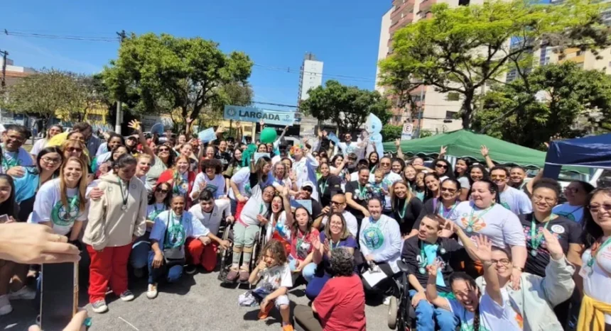 Uma grande foto de grupo tirada ao ar livre, em um dia ensolarado, mostrando dezenas de pessoas participando da 2ª Caminhada da Pessoa com Deficiência. O grupo é diverso e inclui pessoas com e sem deficiência, adultos e crianças, alguns em cadeiras de rodas e alguns usando camisetas brancas com um logo estampado.