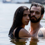 Um plano médio captura um casal, um homem e uma mulher, abraçados na água de um lago ou rio, com a água alcançando aproximadamente o nível do peito. A mulher tem cabelos longos e escuros, vestindo uma peça de roupa preta, e está apoiada no ombro do homem, olhando diretamente para a frente com uma expressão séria. O homem, com barba e cabelo escuro, está com o torso nu, exibindo uma tatuagem no peito/ombro esquerdo, e olha diretamente para a câmera com uma expressão intensa. A luz é natural e brilhante. O fundo é a superfície da água com alguns objetos e terra embaçados ao longe.