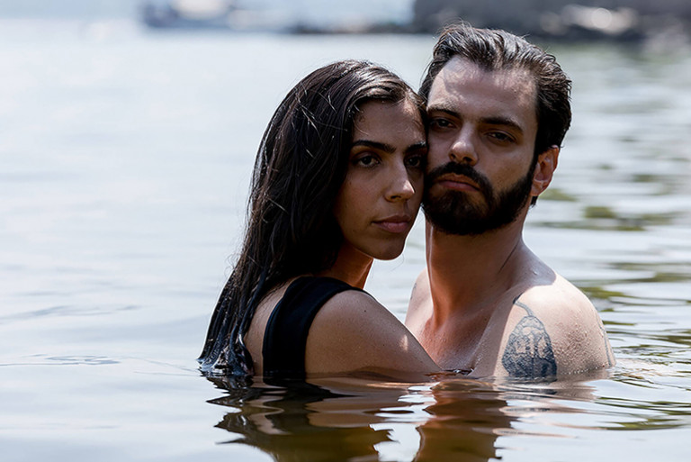 Um plano médio captura um casal, um homem e uma mulher, abraçados na água de um lago ou rio, com a água alcançando aproximadamente o nível do peito. A mulher tem cabelos longos e escuros, vestindo uma peça de roupa preta, e está apoiada no ombro do homem, olhando diretamente para a frente com uma expressão séria. O homem, com barba e cabelo escuro, está com o torso nu, exibindo uma tatuagem no peito/ombro esquerdo, e olha diretamente para a câmera com uma expressão intensa. A luz é natural e brilhante. O fundo é a superfície da água com alguns objetos e terra embaçados ao longe.