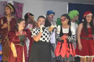 Um grupo diverso de jovens e adultos em trajes de apresentação, em um palco. No centro, uma jovem com síndrome de Down canta em um microfone, vestindo uma blusa de bolinhas e uma saia preta. Ao redor dela, há outras pessoas com e sem deficiência, vestidas como um bobo da corte, pirata, personagens de natal e palhaços.