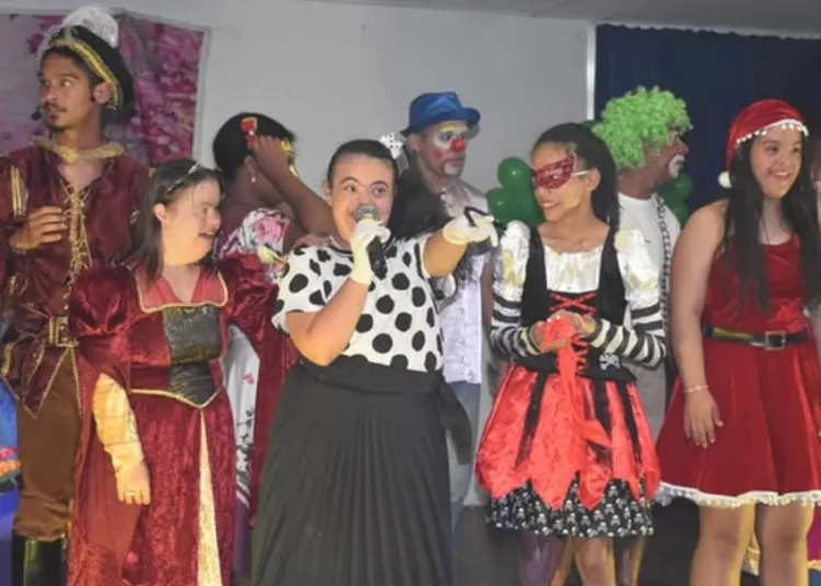 Um grupo diverso de jovens e adultos em trajes de apresentação, em um palco. No centro, uma jovem com síndrome de Down canta em um microfone, vestindo uma blusa de bolinhas e uma saia preta. Ao redor dela, há outras pessoas com e sem deficiência, vestidas como um bobo da corte, pirata, personagens de natal e palhaços.