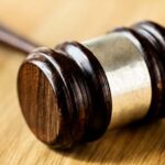 Um martelo de juiz (malhete) de madeira escura com detalhes em metal prateado, repousando inclinado sobre uma mesa de madeira clara. A luz destaca a textura e o brilho do malhete.