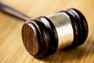 Um martelo de juiz (malhete) de madeira escura com detalhes em metal prateado, repousando inclinado sobre uma mesa de madeira clara. A luz destaca a textura e o brilho do malhete.