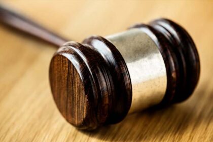 Um martelo de juiz (malhete) de madeira escura com detalhes em metal prateado, repousando inclinado sobre uma mesa de madeira clara. A luz destaca a textura e o brilho do malhete.