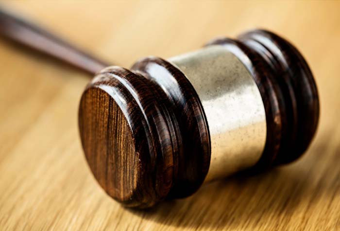 Um martelo de juiz (malhete) de madeira escura com detalhes em metal prateado, repousando inclinado sobre uma mesa de madeira clara. A luz destaca a textura e o brilho do malhete.