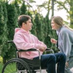 Um jovem sorridente em cadeira de rodas, vestindo um moletom rosa, interage com uma mulher sorridente que está inclinada para ele, segurando uma flor. O casal está ao ar livre, cercado por árvores.