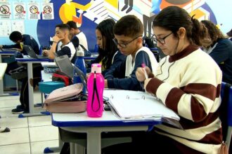 Uma sala de aula com vários alunos sentados em suas carteiras, todos focados em atividades escolares. Em primeiro plano, uma jovem de óculos, cabelo escuro e suéter creme com listras marrons, está concentrada, escrevendo em um caderno de argolas ou ficha. Ela usa fones de ouvido (ou aparelho auditivo) conectados por fio.