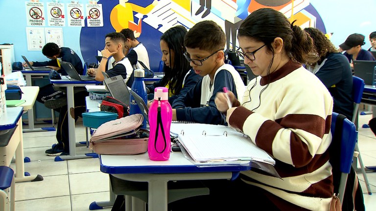 Uma sala de aula com vários alunos sentados em suas carteiras, todos focados em atividades escolares. Em primeiro plano, uma jovem de óculos, cabelo escuro e suéter creme com listras marrons, está concentrada, escrevendo em um caderno de argolas ou ficha. Ela usa fones de ouvido (ou aparelho auditivo) conectados por fio.