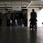 Silhueta de uma pessoa em uma cadeira de rodas sendo empurrada por outra, caminhando por um túnel iluminado em direção a uma área com escadas rolantes e a placa de 'Metrô'. Há outras pessoas ao fundo caminhando na direção oposta.