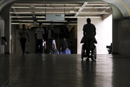Silhueta de uma pessoa em uma cadeira de rodas sendo empurrada por outra, caminhando por um túnel iluminado em direção a uma área com escadas rolantes e a placa de 'Metrô'. Há outras pessoas ao fundo caminhando na direção oposta.
