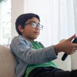 Um close-up lateral mostra um menino jovem, de óculos, sentado em um sofá claro (aparentemente cinza ou bege) em uma sala bem iluminada. O menino, com cabelo escuro, está vestindo uma camisa verde e uma jaqueta jeans azul clara. Ele está segurando um controle remoto preto com a mão direita esticada