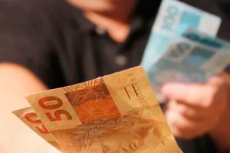 Plano fechado mostra mãos segurando cédulas de reais. Em primeiro plano, notas de R$ 50 são visíveis, com uma nota de R$ 100 mais desfocada sendo segurada ao fundo. A pessoa que segura as notas veste uma camisa preta.