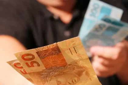 Plano fechado mostra mãos segurando cédulas de reais. Em primeiro plano, notas de R$ 50 são visíveis, com uma nota de R$ 100 mais desfocada sendo segurada ao fundo. A pessoa que segura as notas veste uma camisa preta.