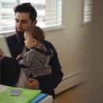 Um homem barbudo, em pé, segura um bebê no colo enquanto olha para um tablet. Eles estão em um ambiente doméstico ou de escritório, com janelas ao fundo.