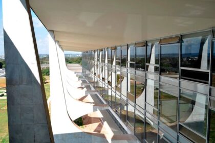 Uma vista em perspectiva da arquitetura moderna de um prédio, possivelmente um edifício governamental em Brasília, como o Palácio do Planalto ou o Palácio do Itamaraty, projetado por Oscar Niemeyer. A imagem mostra o longo corredor de vidro e a faixa de janelas inclinadas que refletem o céu e a paisagem externa.