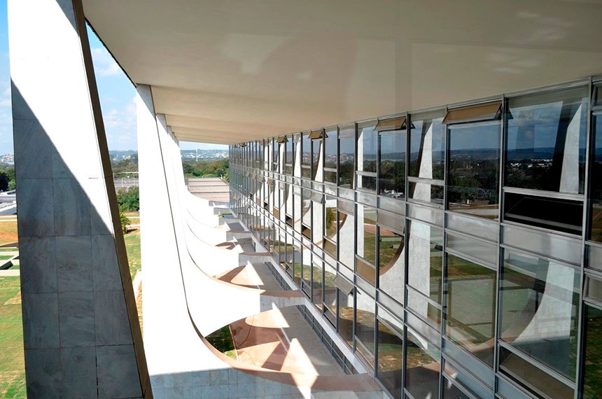 Uma vista em perspectiva da arquitetura moderna de um prédio, possivelmente um edifício governamental em Brasília, como o Palácio do Planalto ou o Palácio do Itamaraty, projetado por Oscar Niemeyer. A imagem mostra o longo corredor de vidro e a faixa de janelas inclinadas que refletem o céu e a paisagem externa.