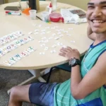 Jovem sorridente sentado à mesa organizando cartões com letras do alfabeto durante uma atividade educativa inclusiva