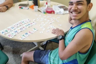 Jovem sorridente sentado à mesa organizando cartões com letras do alfabeto durante uma atividade educativa inclusiva