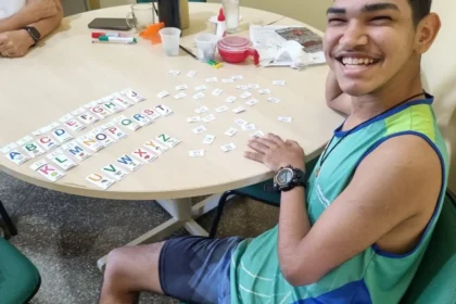 Jovem sorridente sentado à mesa organizando cartões com letras do alfabeto durante uma atividade educativa inclusiva