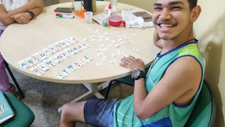 Jovem sorridente sentado à mesa organizando cartões com letras do alfabeto durante uma atividade educativa inclusiva