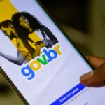 Um close-up de uma mão segurando um smartphone que exibe a tela inicial do aplicativo Gov.br, com o logotipo em destaque e a opção "Entrar com gov.br". O fundo da tela é dividido em cores amarela e preta, com imagens de um homem e uma mulher. Gov.br mais acessível