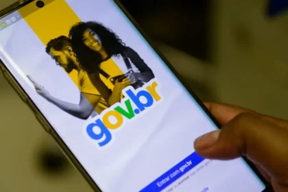 Um close-up de uma mão segurando um smartphone que exibe a tela inicial do aplicativo Gov.br, com o logotipo em destaque e a opção "Entrar com gov.br". O fundo da tela é dividido em cores amarela e preta, com imagens de um homem e uma mulher. Gov.br mais acessível