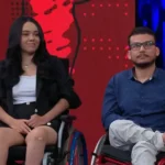 Casal Laila e Luciano, sentado em cadeiras de rodas participa de um programa de TV, com fundo vermelho e azul iluminado.