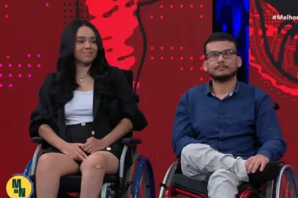 Casal Laila e Luciano, sentado em cadeiras de rodas participa de um programa de TV, com fundo vermelho e azul iluminado.