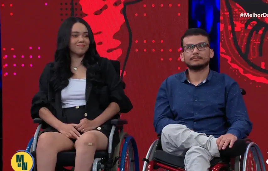 Casal Laila e Luciano, sentado em cadeiras de rodas participa de um programa de TV, com fundo vermelho e azul iluminado.