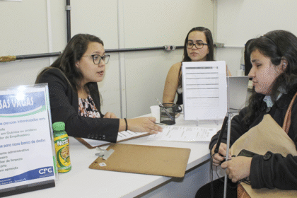 Mulher sendo atendida em um balcão de vagas de emprego, conversando com duas recrutadoras enquanto segura uma bengala.
