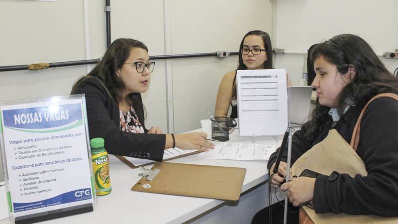 Mulher sendo atendida em um balcão de vagas de emprego, conversando com duas recrutadoras enquanto segura uma bengala.