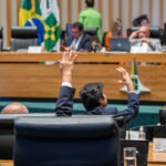 Dois parlamentares levantam as mãos durante uma votação em um plenário, com mesa diretora e bandeiras ao fundo.