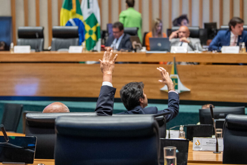 Dois parlamentares levantam as mãos durante uma votação em um plenário, com mesa diretora e bandeiras ao fundo.