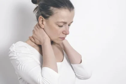 Mulher com expressão de dor segurando o pescoço com as duas mãos, em fundo branco.