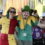 Bloco Deficiente é a Mãe - Duas pessoas fantasiadas e sorrindo, com os braços abertos, em um ambiente festivo decorado com fitas coloridas. Ao fundo, outras pessoas participam da celebração, algumas sentadas em cadeiras plásticas.