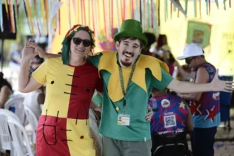 Bloco Deficiente é a Mãe - Duas pessoas fantasiadas e sorrindo, com os braços abertos, em um ambiente festivo decorado com fitas coloridas. Ao fundo, outras pessoas participam da celebração, algumas sentadas em cadeiras plásticas.